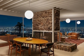 Una mesa de madera con sillas, una chimenea de piedra, sillones y lámparas esféricas están frente a un ventanal panorámico con luces nocturnas