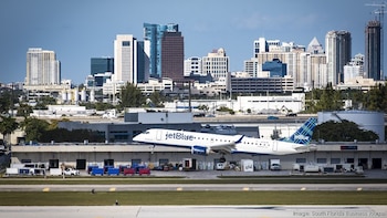 Un avión blanco de JetBlue despega de una pista de aeropuerto, con edificios de la ciudad de Fort Lauderdale y cielo azul de fondo