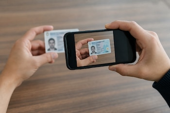 Dos manos sostienen un teléfono móvil cuya pantalla encuadra un documento de identidad que otra mano sujeta sobre una mesa.