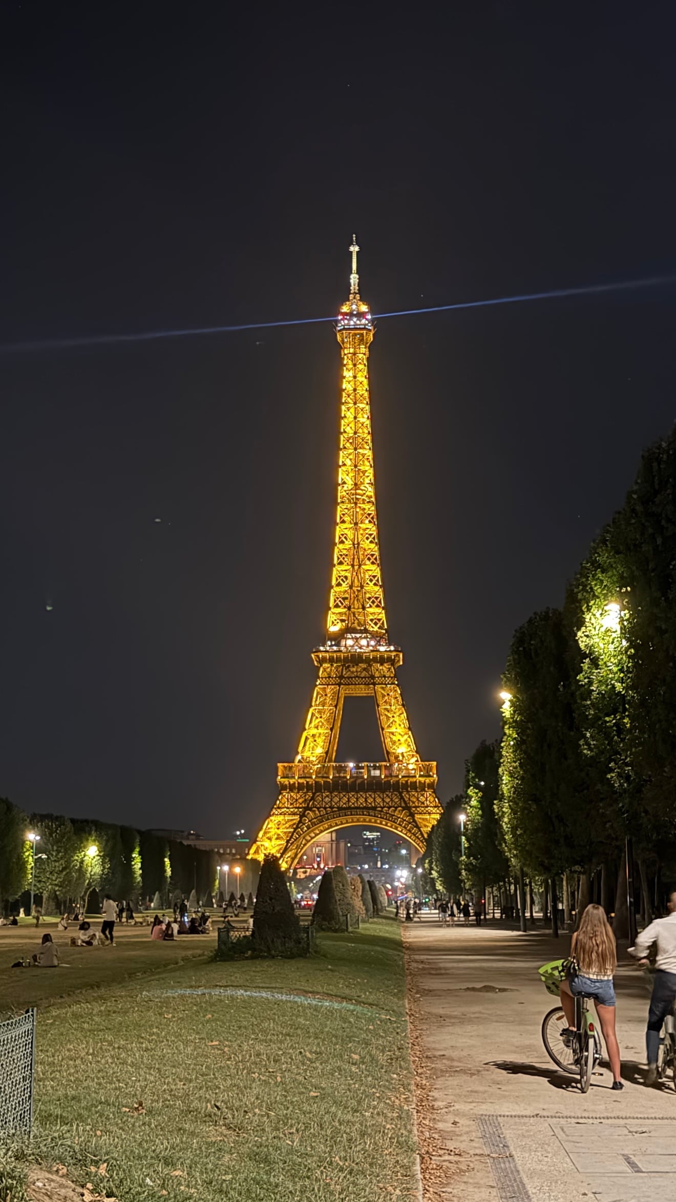 La Torre Eiffel ilumina la noche parisina, mientras personas disfrutan en los jardines y dos ciclistas transitan por el sendero adyacente.