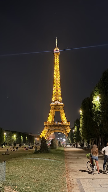 Torre Eiffel dorada de noche. Cielo oscuro. Césped, árboles, farolas. Personas sentadas y caminando en jardines. Dos ciclistas en sendero