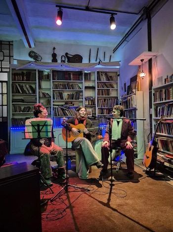 Tres personas están sentadas en una sala llena de libros; la del centro toca una guitarra acústica con un micrófono enfrente