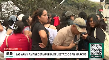 Un grupo de personas se aglomera en una calle junto a árboles y rejas, con un faldón informativo de noticias en la parte inferior