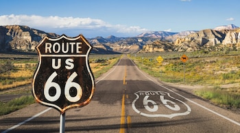 Un cartel oxidado de la Ruta 66 en el lado izquierdo de una carretera desértica con el logo "ROUTE 66" pintado en el asfalto. Al fondo, montañas y cielo azul.