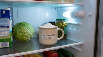 Una taza blanca con bicarbonato de sodio en el estante superior de un refrigerador iluminado, junto a lechuga, un frasco de pepinillos y leche.