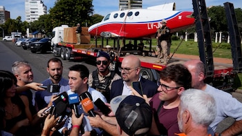 Avión blanco y rojo sobre camión de plataforma, custodiado por seguridad, rodeado de periodistas en calle urbana