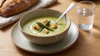 Un plato hondo con sopa verde y cuchara. Contiene puntas de espárrago, croutones y crema. Al lado hay un vaso con agua, un pan y un mantel sobre mesa de madera.