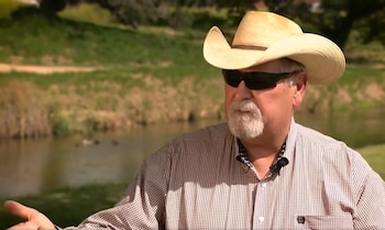 Un hombre de mediana edad con sombrero de vaquero claro, gafas de sol oscuras, barba y camisa a cuadros, está sentado frente a un río y vegetación