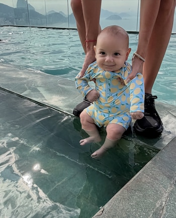 Un bebé sonriendo en una piscina, sostenido por manos de adulto. Usa un traje de baño azul claro con limones amarillos. De fondo se ven montañas y edificios