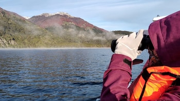 Persona con chaqueta morada y chaleco salvavidas que observa con binoculares un lago rodeado de montañas con neblina