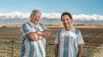 Dos hombres, uno de cabello gris y otro de cabello rizado oscuro, sonríen en camisetas de Argentina frente a un viñedo y montañas nevadas bajo un cielo azul.