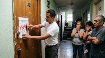 Un hombre con camiseta blanca sella un papel en una puerta de madera ante tres personas en el pasillo de un edificio.
