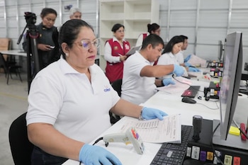 Un grupo de personas, incluyendo una mujer en primer plano con gafas y guantes azules, trabaja en escritorios escaneando documentos y utilizando computadoras