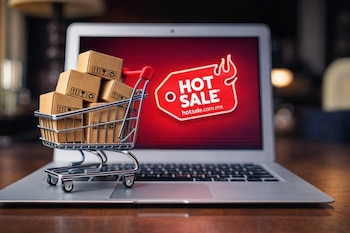 Un carrito de compras en miniatura lleno de cajas de cartón sobre el teclado de una laptop. La pantalla muestra el logo rojo de Hot Sale.