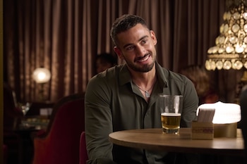 Nicolás Occhiato viste camisa verde y sonríe sentado junto a una mesa de madera con un vaso de cerveza en un espacio iluminado con luz cálida
