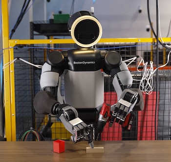 Robot humanoide blanco y negro de Boston Dynamics con un taladro rojo en la mano sobre una mesa junto a una pieza de madera y un recipiente rojo