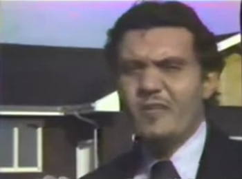 Primer plano de un hombre con cabello oscuro y bigote, vistiendo traje y corbata, con el tejado y la canaleta de una casa visible al fondo