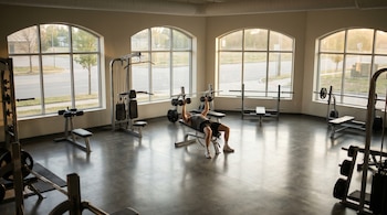 Hombre en camiseta y shorts entrena con mancuernas en un banco de pesas. El gimnasio está vacío, con grandes ventanales y luz natural matutina.