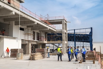Edificio en construcción con estructuras de concreto y acero. Trabajadores con cascos y chalecos de seguridad observan el sitio, con andamios y materiales apilados