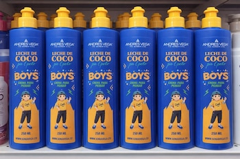 Estante con múltiples botellas azules de crema con tapa amarilla. Las etiquetas muestran "Leche de Coco pa'l pelo Boys" de Andres Vega y un niño.