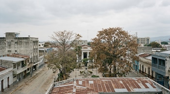 Vista aérea de una plaza y calles secas en San Salvador bajo un cielo nublado. Se observan edificios y árboles urbanos con follaje marchito.