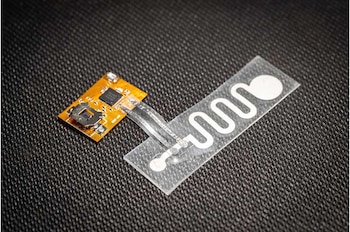 Módulo electrónico con placa de circuito impreso naranja, componentes, y tira flexible transparente con un patrón de circuito blanco sobre fondo oscuro