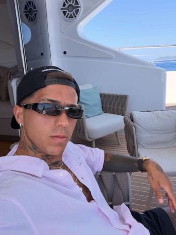 Un hombre con gorra negra, gafas de sol oscuras y camisa rosa clara, sentado en la cubierta de un yate. Luce tatuajes en el cuello y los brazos