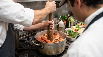 Las manos de un cocinero sostienen un mazo de madera para triturar cáscaras de bogavante en una olla sobre una estufa.