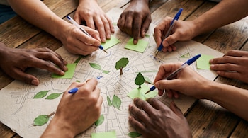 Manos de varias personas marcan un mapa urbano con bolígrafos, notas adhesivas verdes y figuras de árboles de papel sobre una mesa de madera.