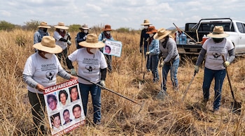 Grupo de personas con sombreros y mascarillas rastrea un campo de pasto seco con herramientas. Algunos sostienen carteles con fotos de desaparecidos.