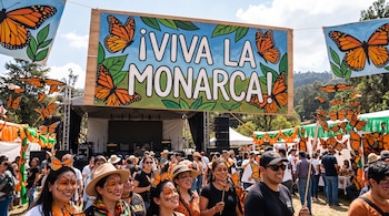 Un cartel con el texto Viva la Monarca sobre personas con pintura facial, figuras de mariposas colgantes y un escenario al aire libre.