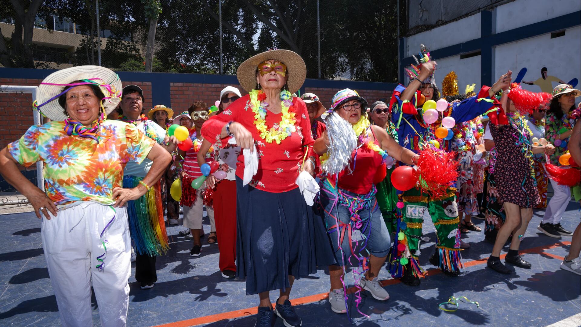 Un grupo de adultos mayores baila y celebra con vestimentas coloridas, globos y confeti en una cancha al aire libre, conmemorando el Día de San Valentín y la renovación de sus votos matrimoniales.