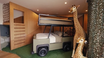 Dormitorio infantil con cama cucheta tipo vehículo safari, dos jirafas de peluche, suelo de césped artificial, pared de madera y red de escalada