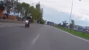 Imagen de un motociclista con casco y chaqueta oscura en una autopista con múltiples carriles, rodeado de vegetación y postes de luz