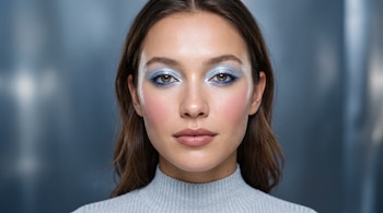 Retrato de una mujer con sombra de ojos azul perlada, labios rosa y suéter gris de cuello alto ante un fondo azul.
