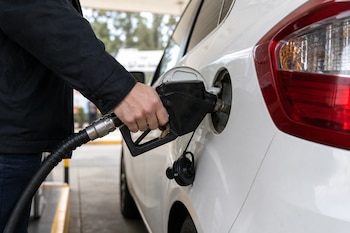 Una mano con chaqueta oscura sostiene la manguera del surtidor para cargar combustible en un vehículo blanco.