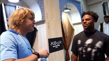 Primer plano de dos hombres, uno rubio en camiseta azul, el otro moreno en camiseta negra, conversando en un espacio con arcos y un cartel en árabe