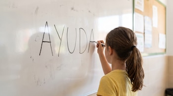 Niña de espalda con camiseta amarilla y coleta escribe la palabra AYUDA en un tablero blanco con un marcador negro en un aula escolar.