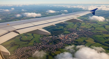 Ala de avión en primer plano sobrevolando campos verdes, ciudades pequeñas y nubes. El horizonte muestra un cielo despejado con nubes dispersas.
