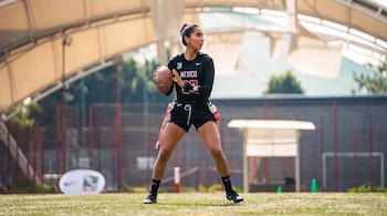Una mujer vestida con uniforme de flag football de México, camiseta negra con número 33 y shorts, sostiene un balón de fútbol americano en un campo.