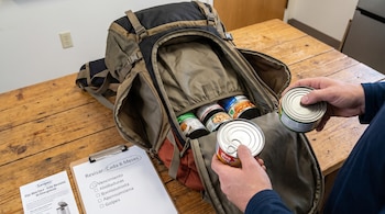Manos de persona colocando latas de conservas en una mochila abierta. En la mesa, hay varias latas, un portapapeles con lista de revisión y un folleto de Sanipes