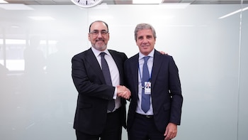 Luis Caputo y Sergio Diaz-Granados, ambos en traje, se dan la mano sonriendo frente a una pared blanca y un reloj de pared