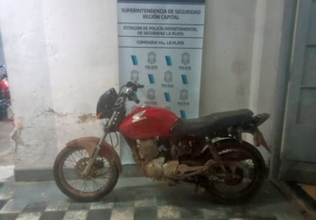 Motocicleta Honda de color rojo estacionada frente a un cartel de la Superintendencia de Seguridad Región Capital y Comisaría 14ta. La Plata