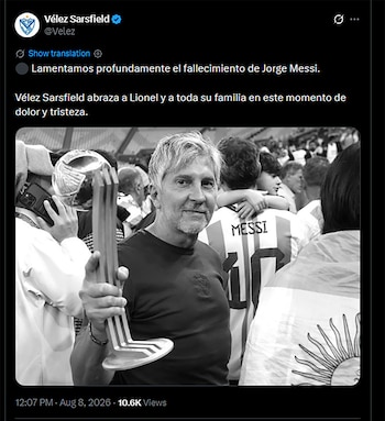 Hombre canoso y barbudo, con camiseta oscura, sostiene un trofeo. Detrás, personas; una viste camiseta con 'Messi 10', otra tiene bandera argentina