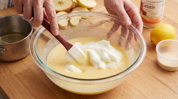 Manos mezclan en un bol de vidrio una crema amarilla con claras batidas. Se observan manzanas en rodajas, una olla, un limón y un envase de gelatina en polvo.