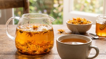 Tetera de vidrio con infusión humeante y flores, taza servida, tazón con flores secas y tarro de miel sobre mesa de madera.