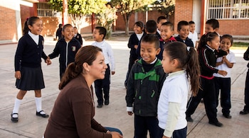 Una mujer agachada habla con una niña y un niño en el patio de una escuela rodeada por varios alumnos con uniformes escolares.