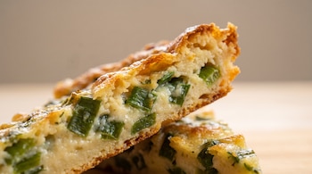 Un trozo de fainá con cebolla de verdeo cortado en diagonal, con bordes dorados y un interior cremoso con trozos de cebolla verde.