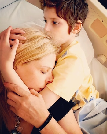 Una mujer rubia acostada en una cama abraza a un niño con camiseta amarilla que sostiene su cabeza