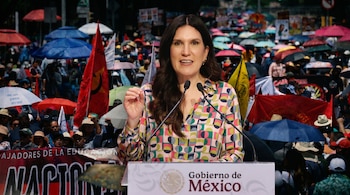 Mujer con cabello oscuro y blusa colorida habla en un podio del Gobierno de México. Detrás, una multitud de personas con sombrillas rojas y azules.